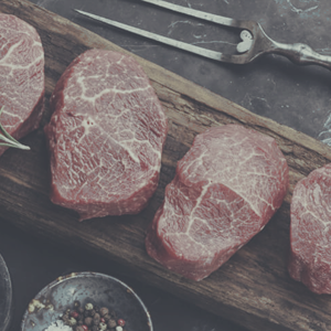 A wooden board with four raw steak on it.